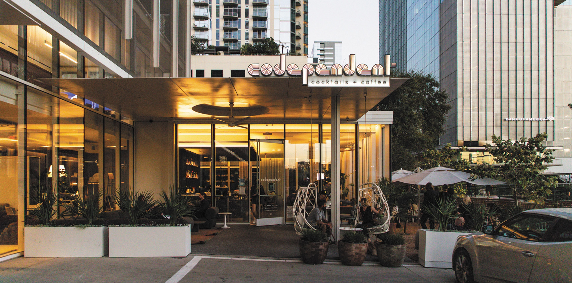 The Codependent entrance at dusk, its neon sign lit, with hanging chairs and agave planters below the downtown towers.
