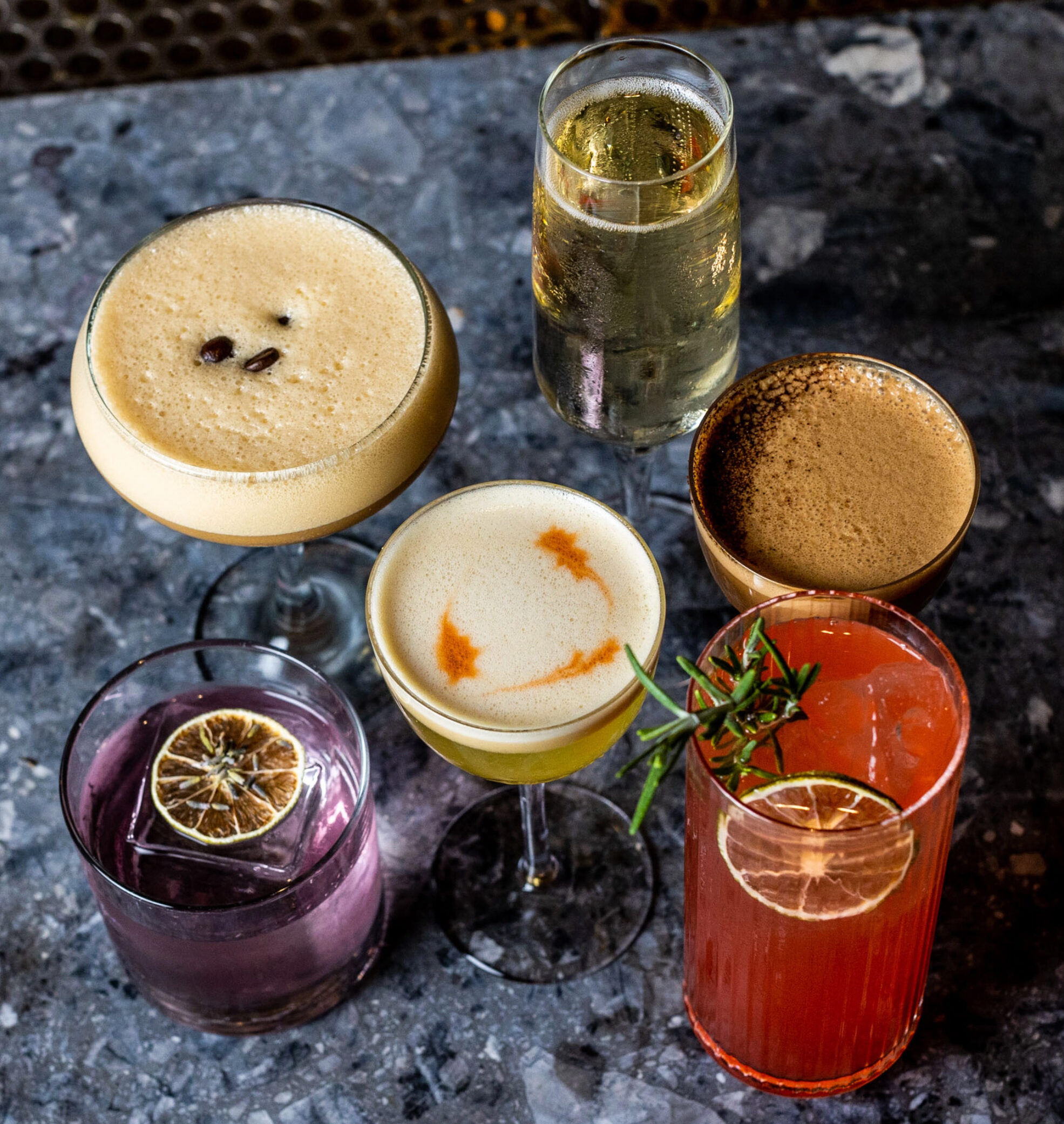 Six cocktails on a dark terrazzo bar top, including an espresso martini, a wine flute and a tall red drink with rosemary.