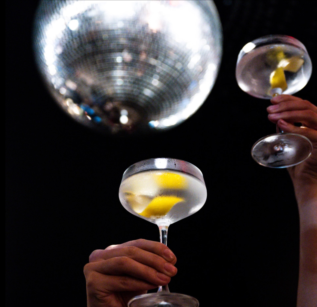 Two hands raising martini coupes with lemon twists beneath a mirrorball.