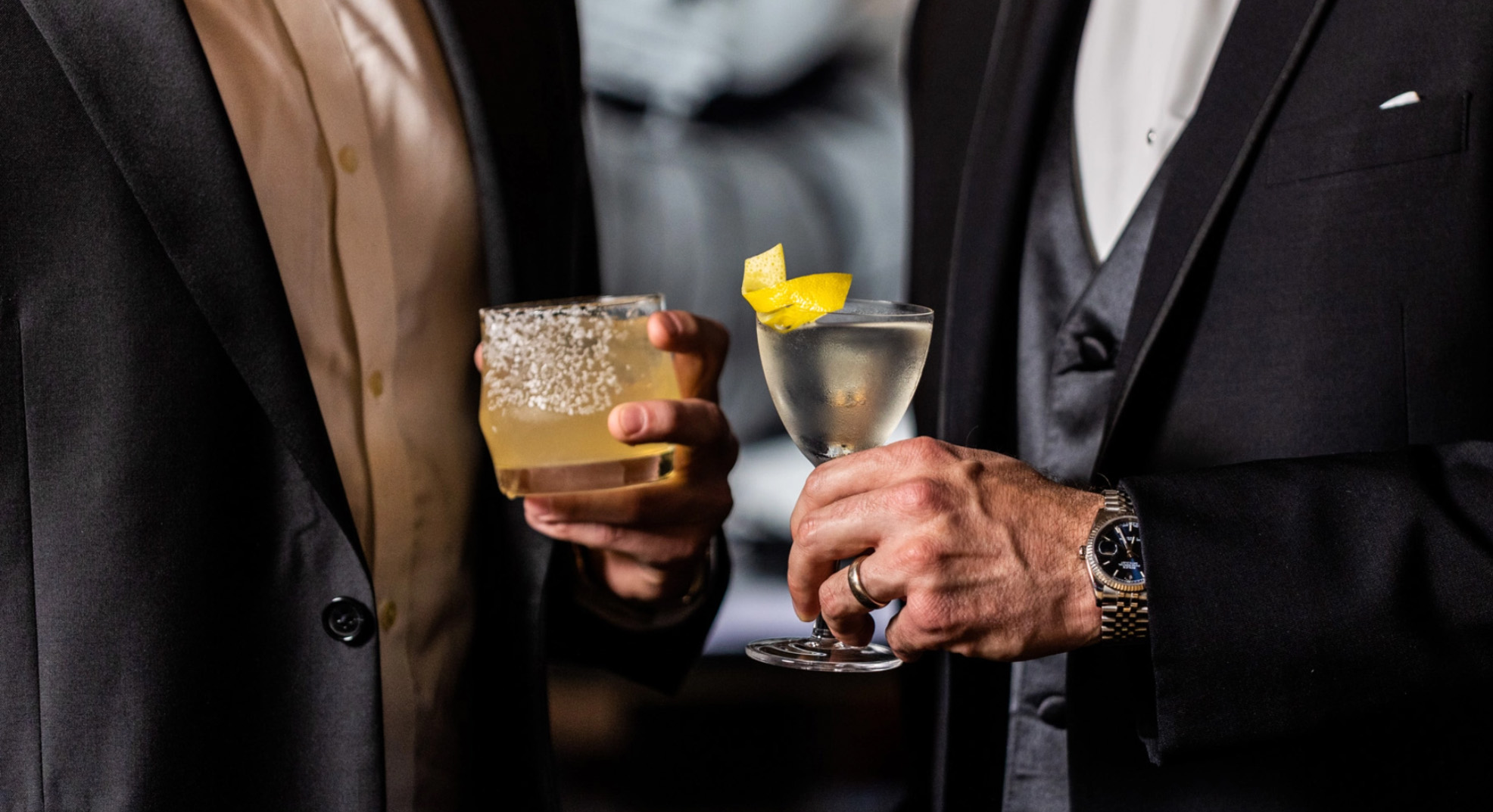 Two guests in evening dress holding a salt-rimmed rocks cocktail and a martini garnished with a lemon twist.