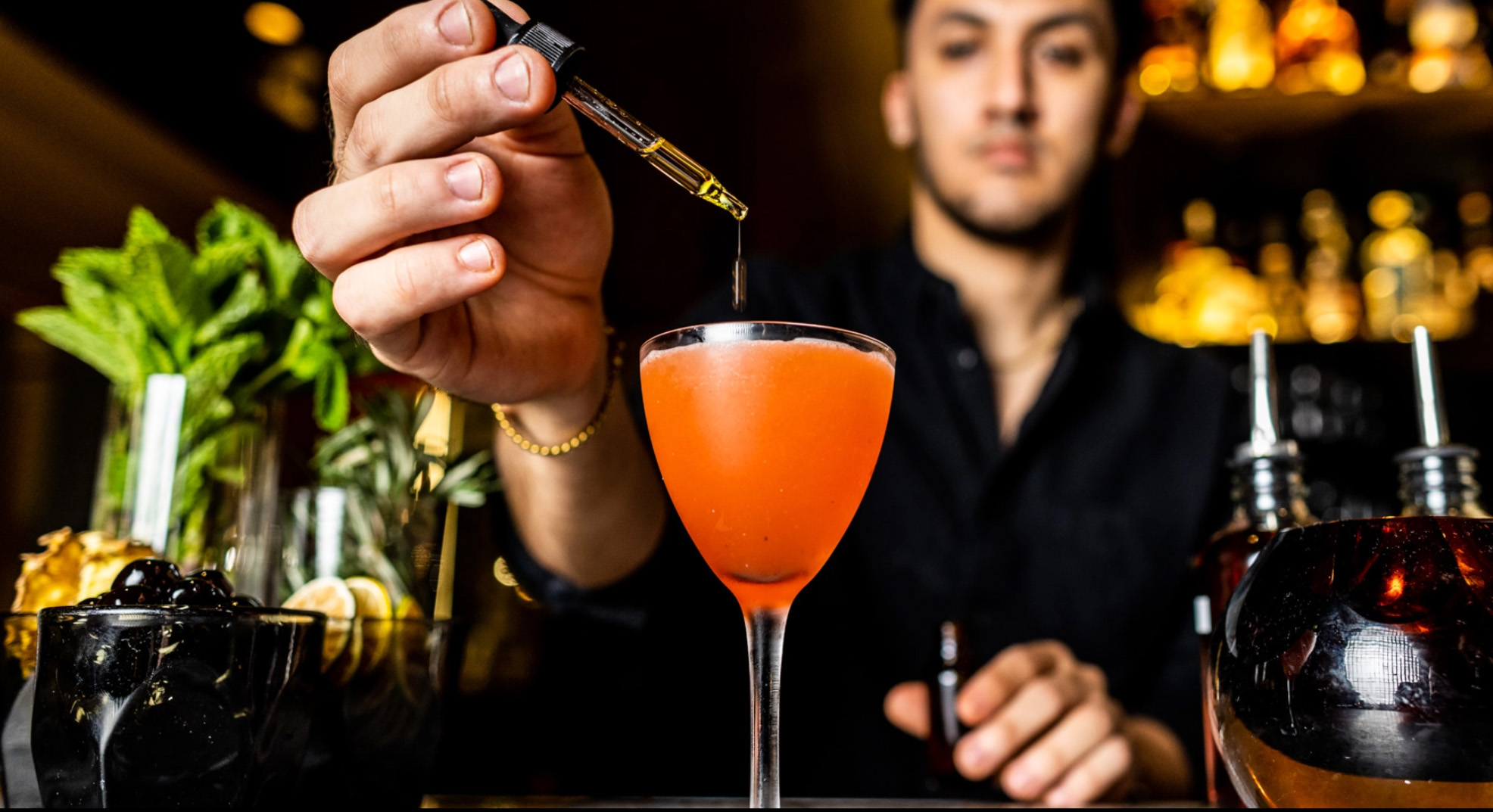 A bartender finishing a coral coloured cocktail with a dropper of bitters, the backlit spirits shelf behind them.