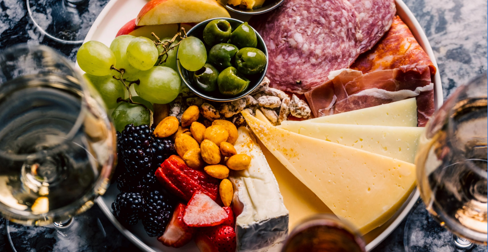 A charcuterie board of salami, prosciutto, sliced cheeses, olives, grapes, berries and almonds, surrounded by wine glasses.