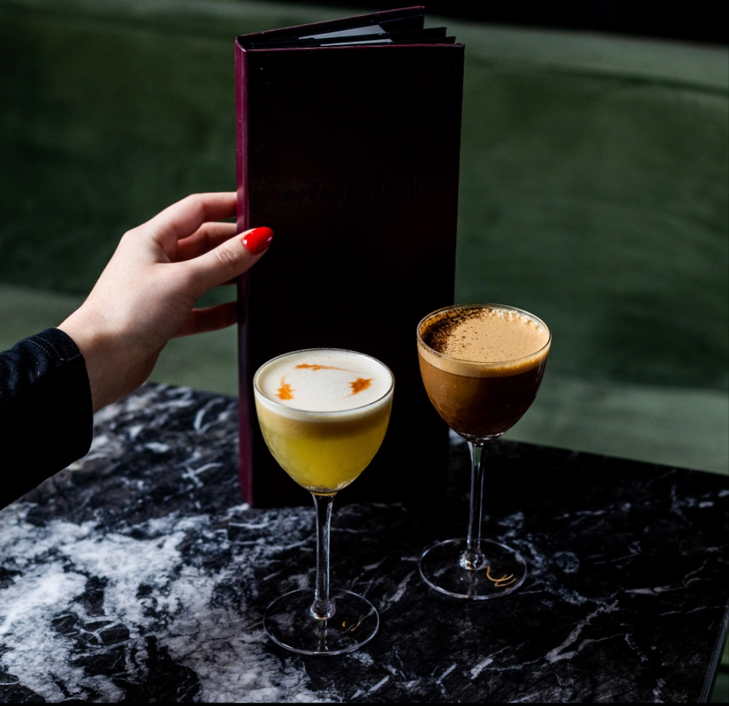 A guest holding the Codependent menu beside two coupe cocktails on black marble, one dusted and one finished with bitters.