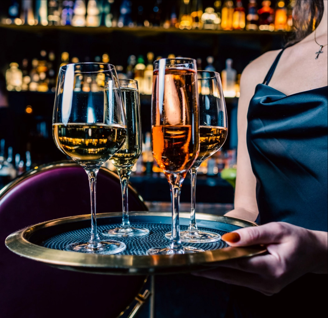 A server carrying a brass tray of white, sparkling and rose wine past the lit back bar.